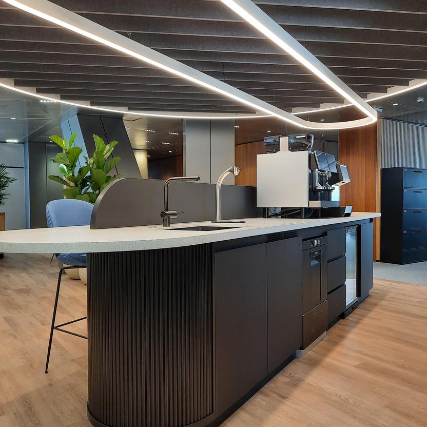 Eine schicke Büroküche mit geschwungenen LED-Leuchten, dunklen Schränken und einer Espressomaschine.