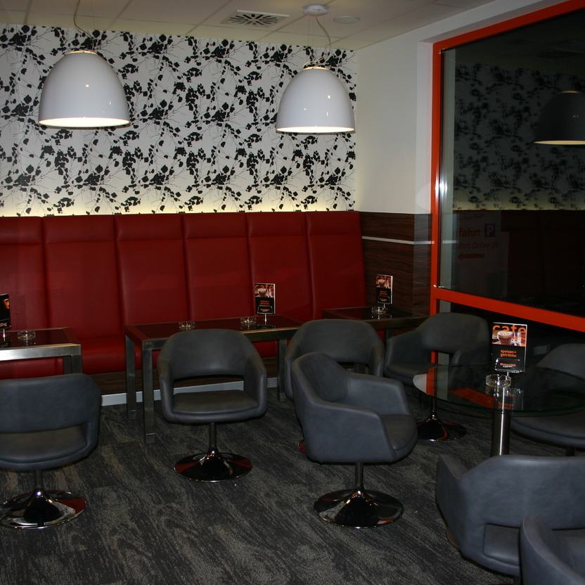 Café-Ecke mit roten Sitzbänken, grauen Sesseln und dekorativer Wandbeleuchtung.