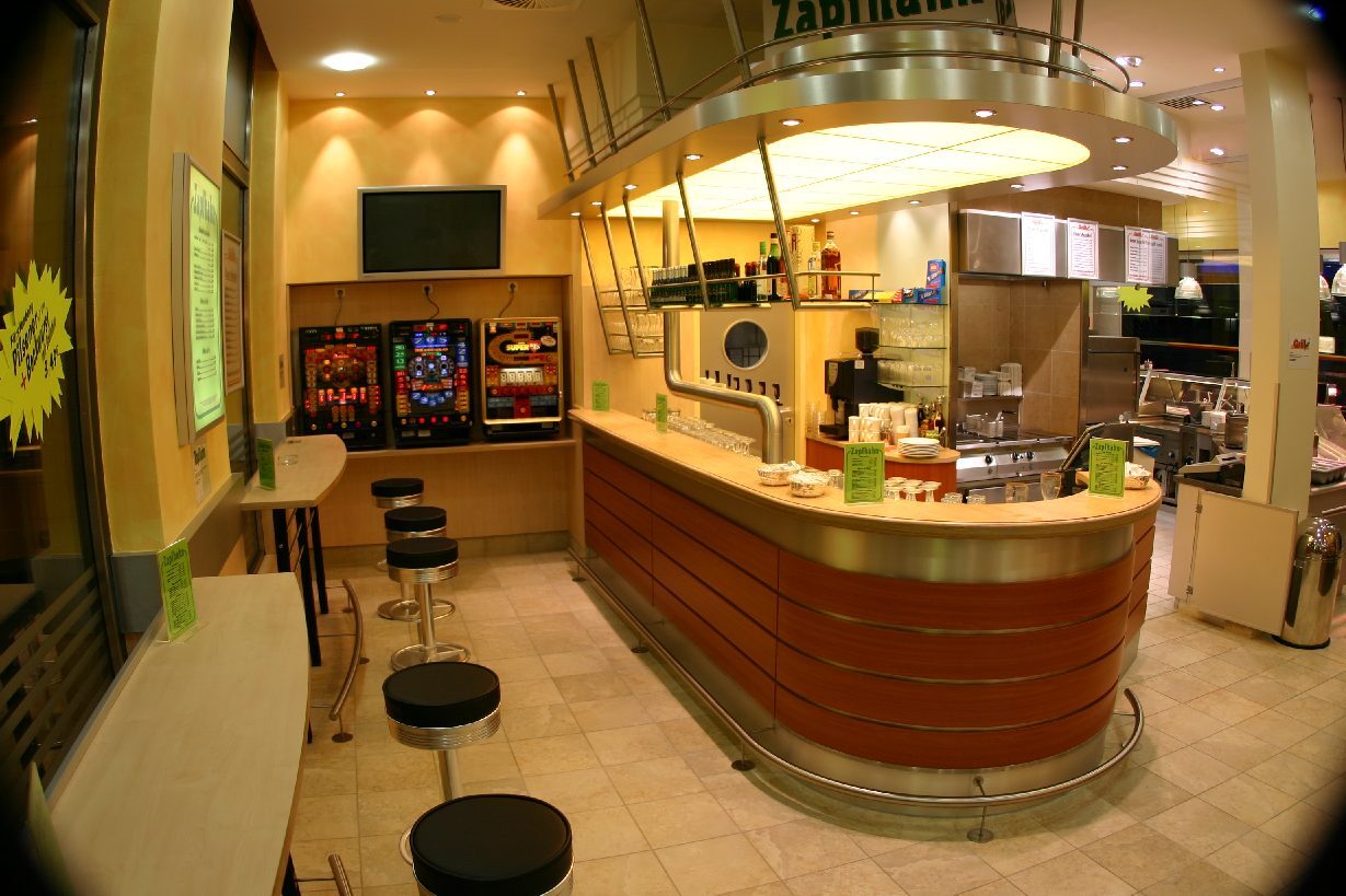 Moderne Caf&eacute;bar mit Holztheke, Sitzpl&auml;tzen und Spielautomaten, beleuchtet mit warmem Licht.