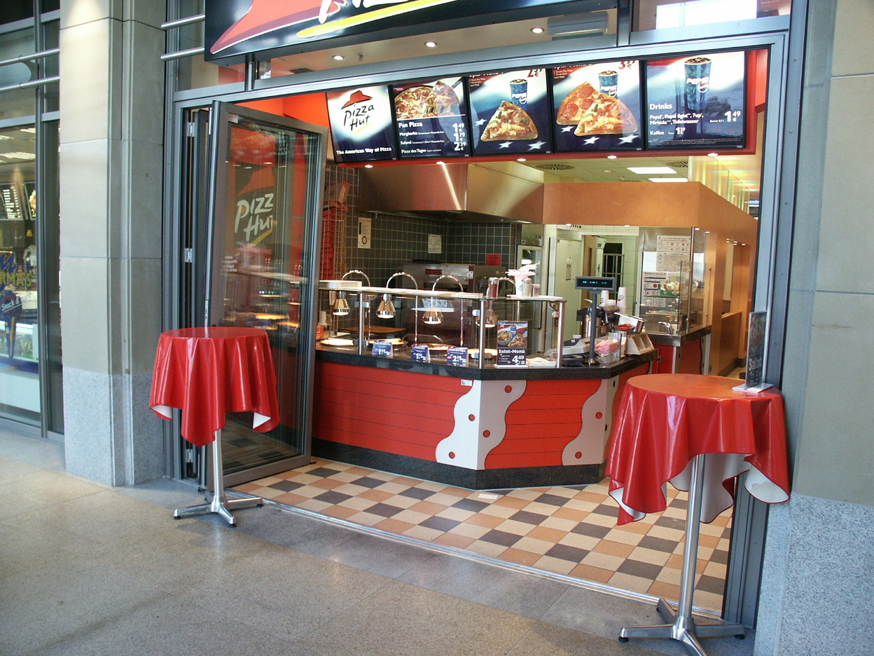 Eingang eines Pizza-Hut-Restaurants mit Theke und roten Tischdecken im Freien. Men&uuml;tafeln &uuml;ber dem Eingang sichtbar.