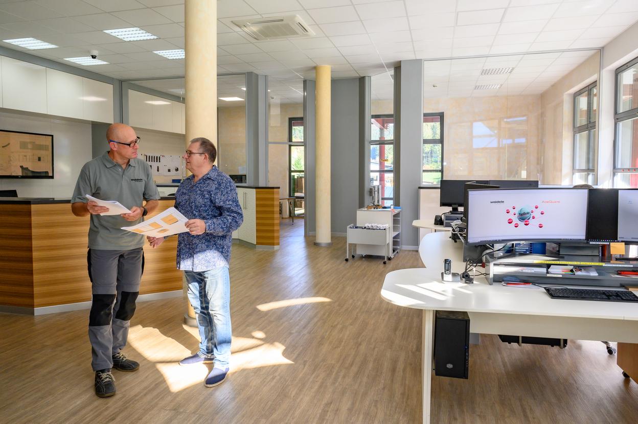 Zwei M&auml;nner stehen in einem modernen B&uuml;roraum und besprechen Dokumente, w&auml;hrend ein Schreibtisch mit Computern im Hintergrund sichtbar ist.