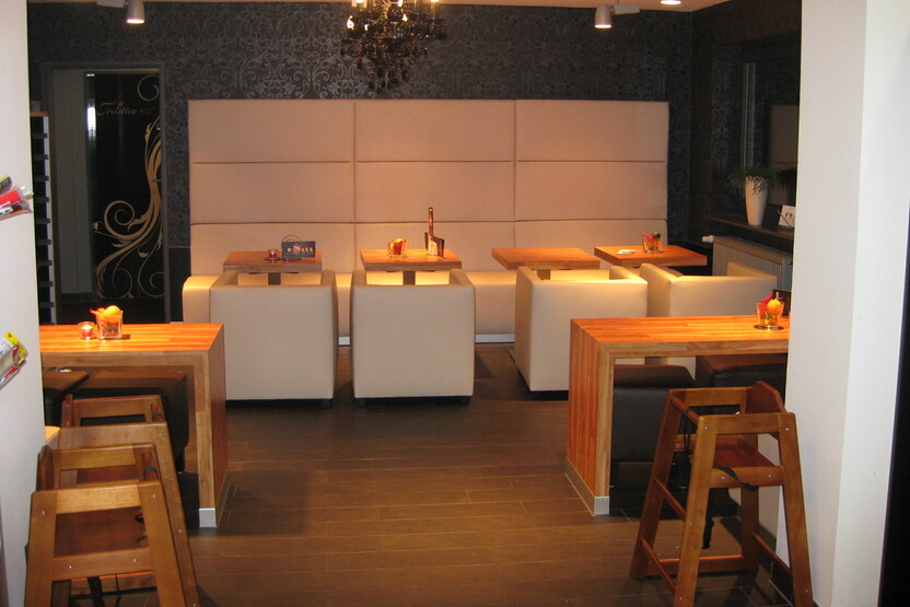 Intimes Ladenbau Gastronomie-Design mit beige gepolsterten Sitzbänken, Holztischen und elegantem Kronleuchter.