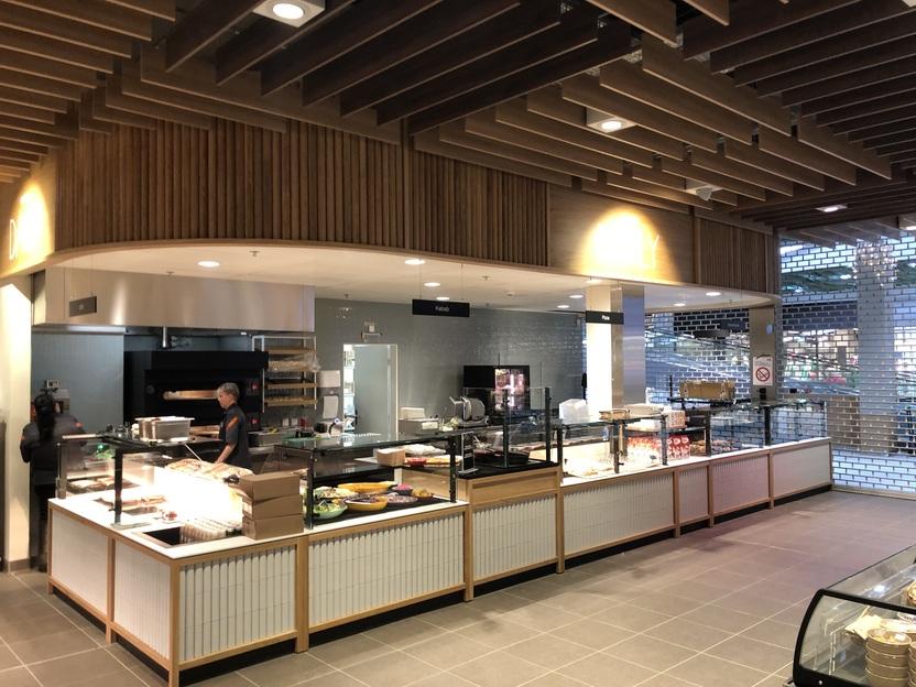 Ein moderner Selbstbedienungsbereich in einer Cafeteria mit Theke, Auslagen und Kochbereich im Hintergrund.